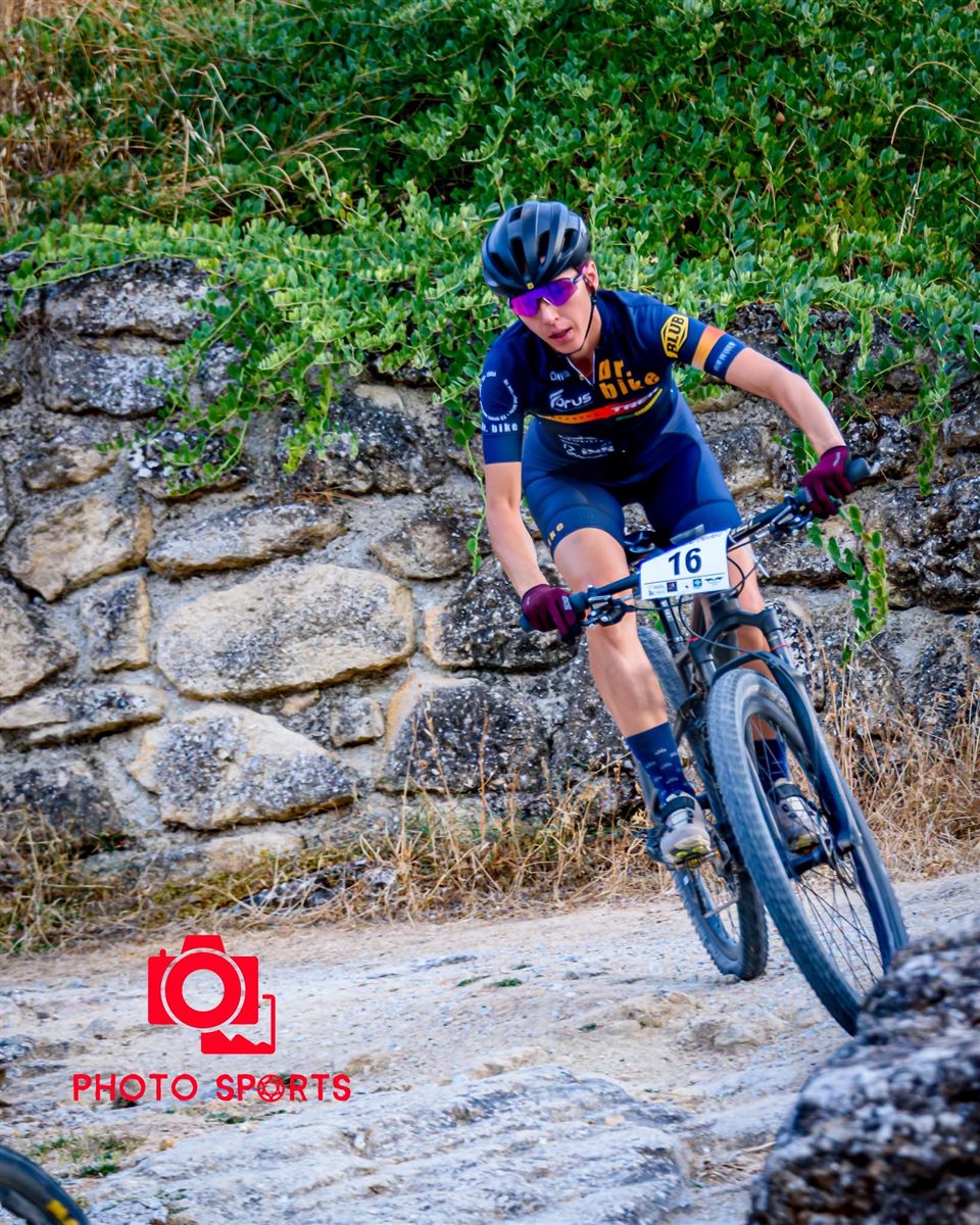 Ana Gay cornering on her mountain bike in front of a dry-stone wall, race number 16